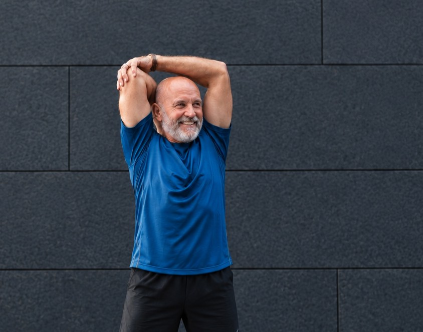 A smiling older man in a blue T-shirt stretches his arms overhead against a dark wall.